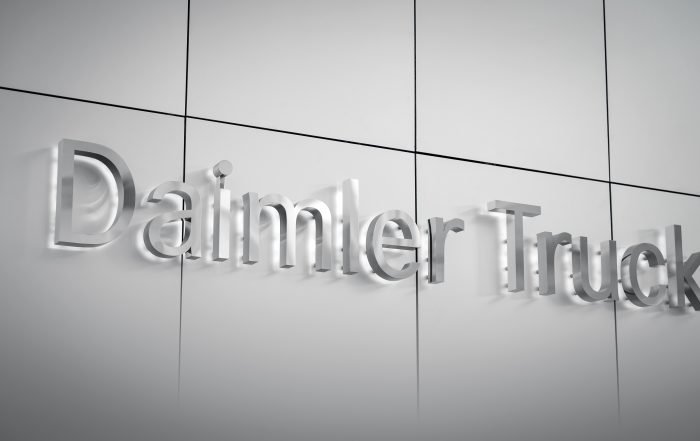 Logo Daimler Truck tridimensionale e metallico su una parete grigio chiaro. Scritta 'Daimler Truck' in rilievo, con ombre che creano profondità. Rappresenta il marchio e la sua presenza nel settore dei veicoli industriali