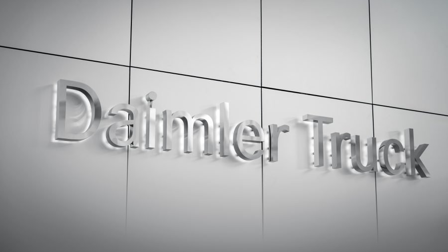 Logo Daimler Truck tridimensionale e metallico su una parete grigio chiaro. Scritta 'Daimler Truck' in rilievo, con ombre che creano profondità. Rappresenta il marchio e la sua presenza nel settore dei veicoli industriali