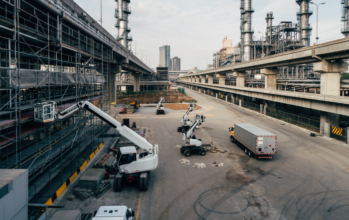 Cantiere industriale con piattaforme di lavoro aeree e veicoli di servizio operativi in un’area infrastrutturale complessa, tra impianti industriali, strutture sopraelevate e ponteggi, in contesto di manutenzione e costruzione.